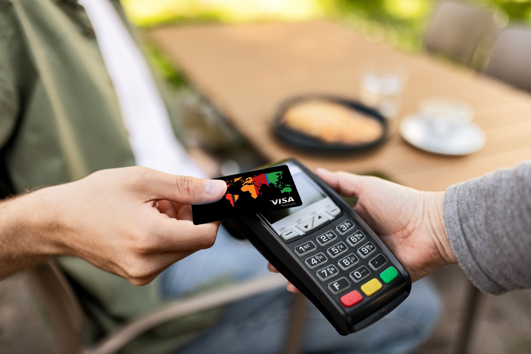Zdjęcie przedstawia płatność zbliżeniową – osoba przykłada kartę płatniczą Visa Świat Intensive z kolorowym motywem mapy świata do terminala trzymanego przez drugą osobę.