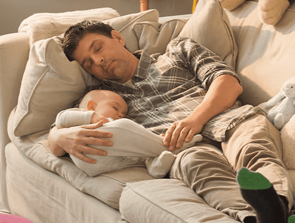 Mężczyzna odpoczywa na kanapie, trzymając śpiące dziecko na rękach — spokojna, domowa scena symbolizująca bezpieczeństwo i spokój w domu.