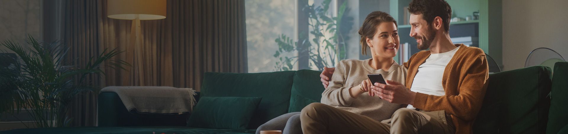 Uśmiechnięta para siedzi razem na kanapie, trzymają telefon. Na grafice napis: 'By się dobrze mieszkało! Ubezpiecz nieruchomość w wygodny dla siebie sposób.' oraz przycisk 'oblicz składkę'