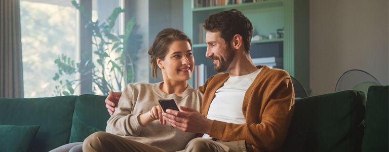 Uśmiechnięta para siedzi razem na kanapie, trzymają telefon. Na grafice napis: 'By się dobrze mieszkało! Ubezpiecz nieruchomość w wygodny dla siebie sposób.' oraz przycisk 'oblicz składkę'