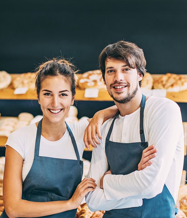 Zdjęcie przedstawia uśmiechniętą kobietę i mężczyznę w fartuchach stojących w piekarni na tle półek z pieczywem.