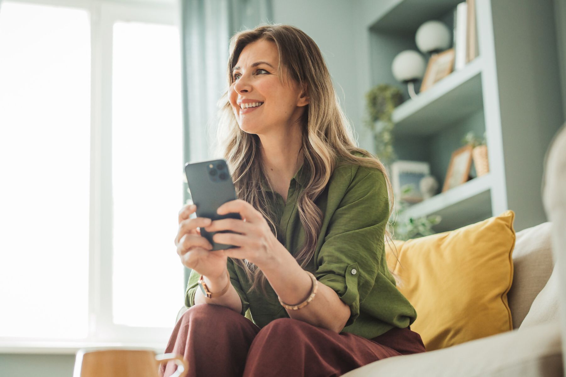 Zdjęcie przedstawia uśmiechniętą kobietę, która siedzi na kanapie i trzyma w dłoniach smartfon.