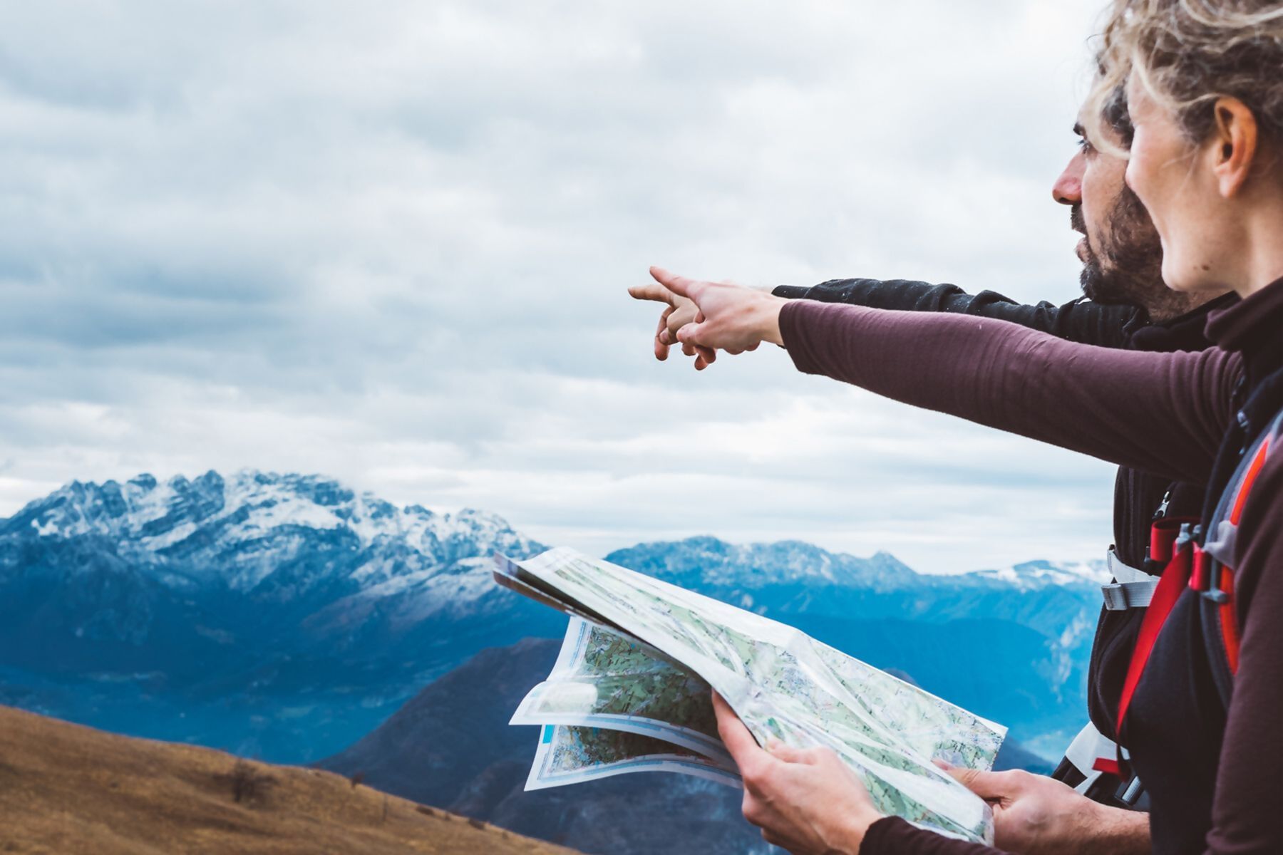 Dwoje turystów, kobieta i mężczyzna, wskazują palcem w dal na tle ośnieżonych gór. Kobieta trzyma w dłoniach mapę.
