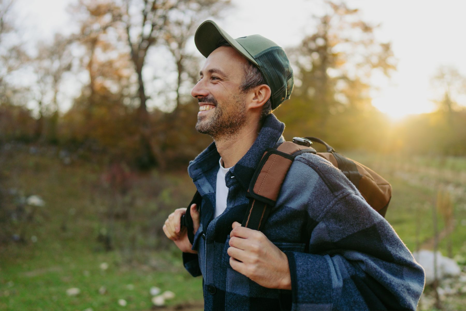 Uśmiechnięty mężczyzna w średnim wieku w czapce z daszkiem i z plecakiem na ramionach, patrzy z radością w dal. Jego twarz oświetla ciepłe, zachodzące słońce, a w tle rozciąga się jesienny krajobraz z drzewami o brązowo-złotych liściach i zieloną roślinnością. Scena emanuje spokojem i pozytywną energią.