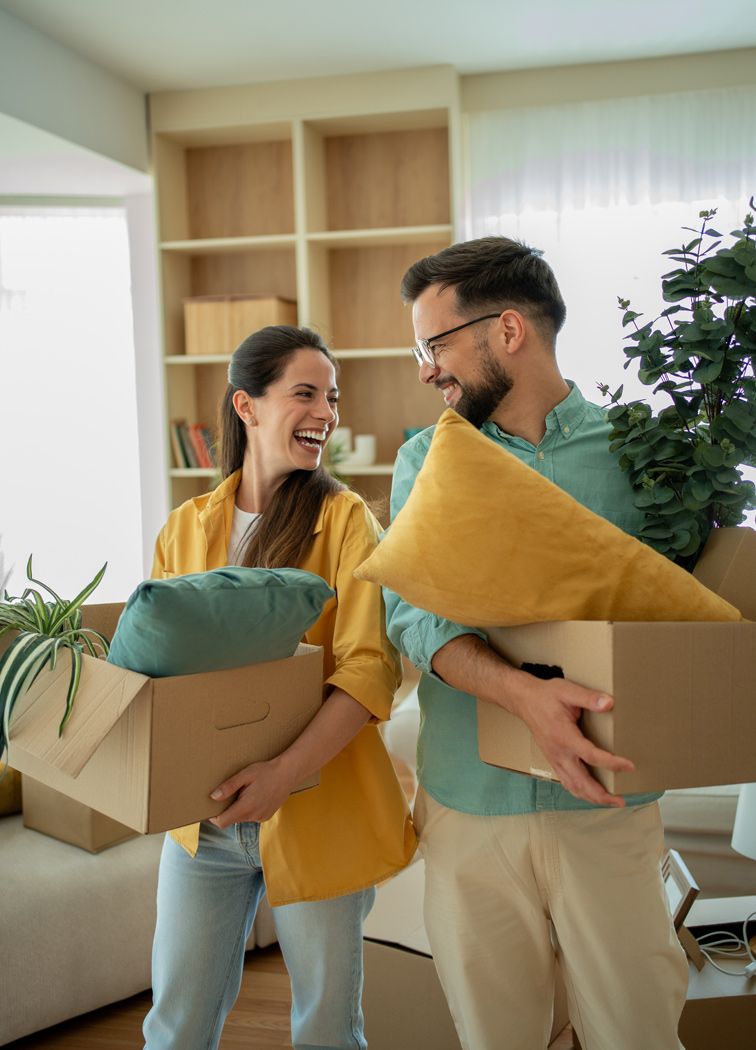 Uśmiechnięta para trzymająca kartony z poduszkami i roślinami podczas przeprowadzki do nowego mieszkania.