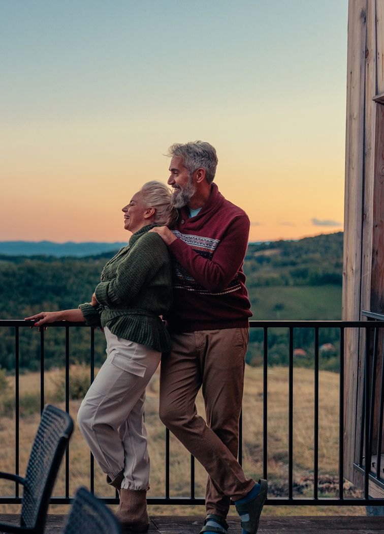 Starsza para obejmująca się i patrząca na zachód słońca z balkonu z widokiem na zielone wzgórza.