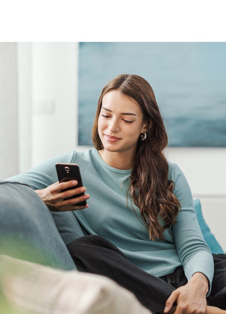 Młoda kobieta siedząca wygodnie na sofie i korzystająca ze smartfona w domowym wnętrzu.