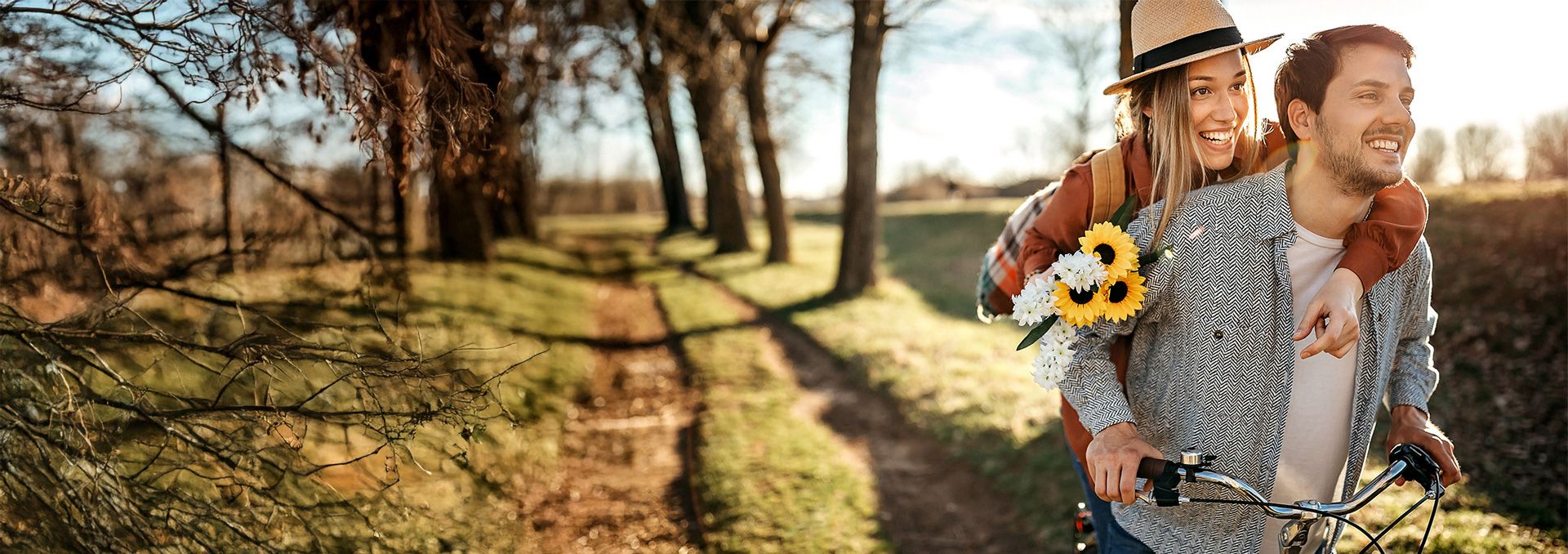 Uśmiechnięta para jedzie rowerem po wiejskiej drodze. Kobieta siedzi z tyłu, obejmuje mężczyznę i trzyma bukiet kwiatów.