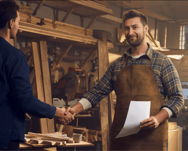 Dwóch mężczyzn w warsztacie stolarskim, stolarz stojący przodem do obiektywu uśmiecha się, trzyma w dłoni kartkę papieru. Widoczne logo mBanku i hasło „Otwórz konto firmowe za 0 zł na zawsze i ciesz się sukcesem we własnej firmie
